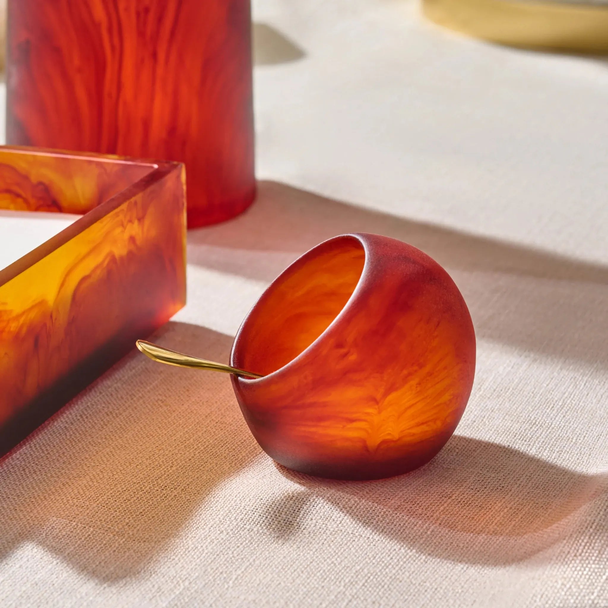 A small rounded amber-orange salt cellar with a gold spoon resting inside, styled on a beige fabric surface with matching amber glassware in the background.