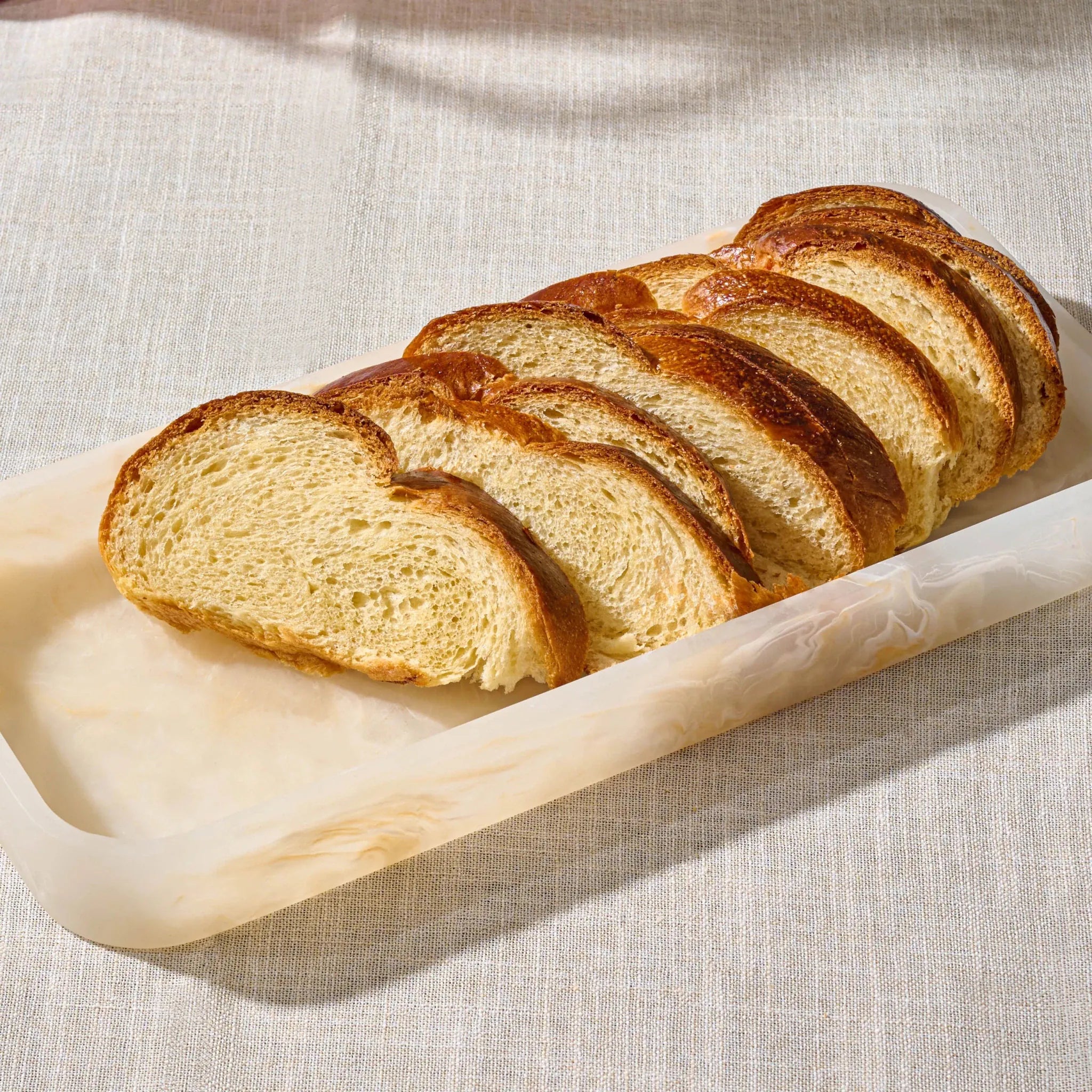 Slices of golden-brown artisan bread arranged diagonally on a translucent rectangular servingresin tray over a textured beige fabric surface.
