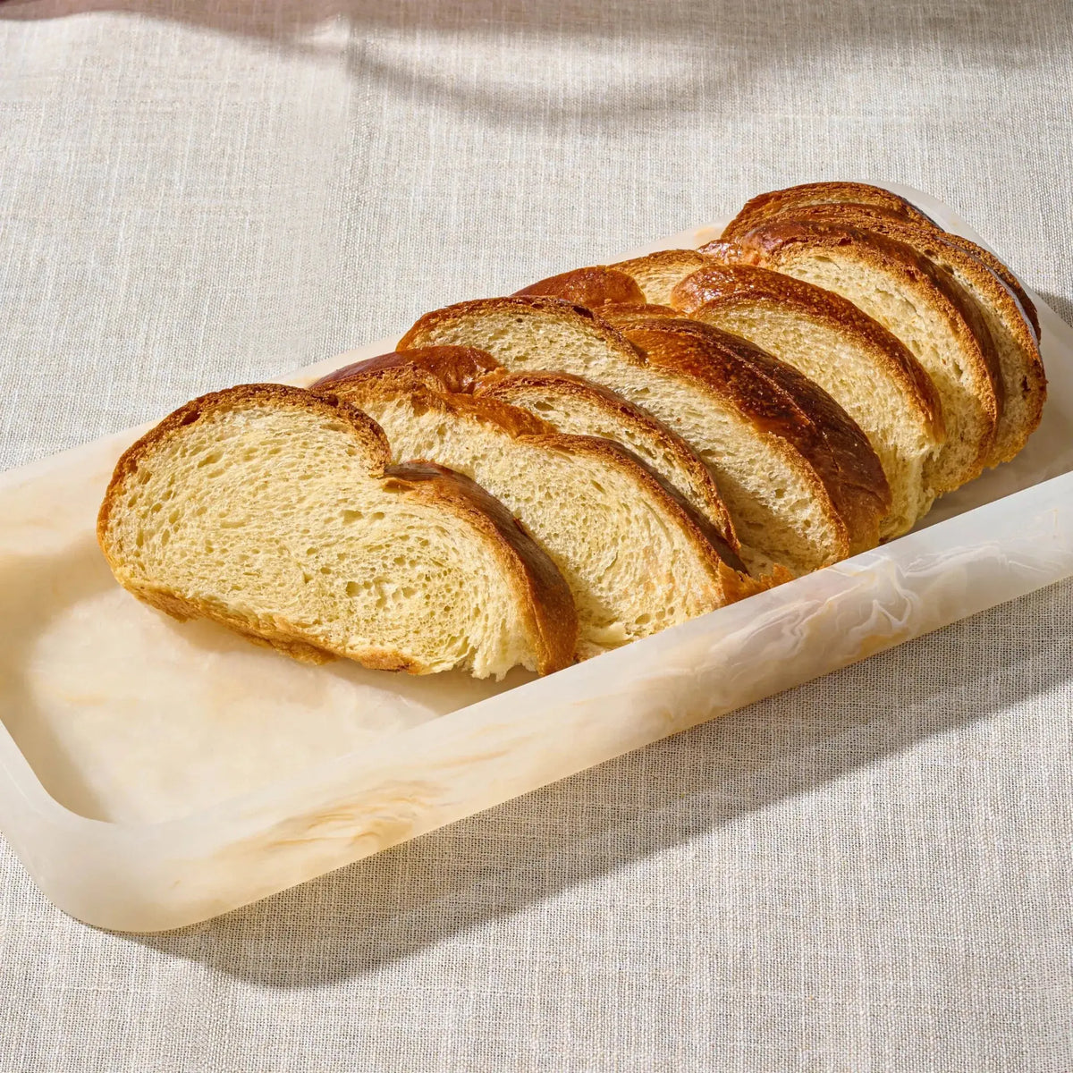 Slices of golden-brown artisan bread arranged diagonally on a translucent rectangular servingresin tray over a textured beige fabric surface.