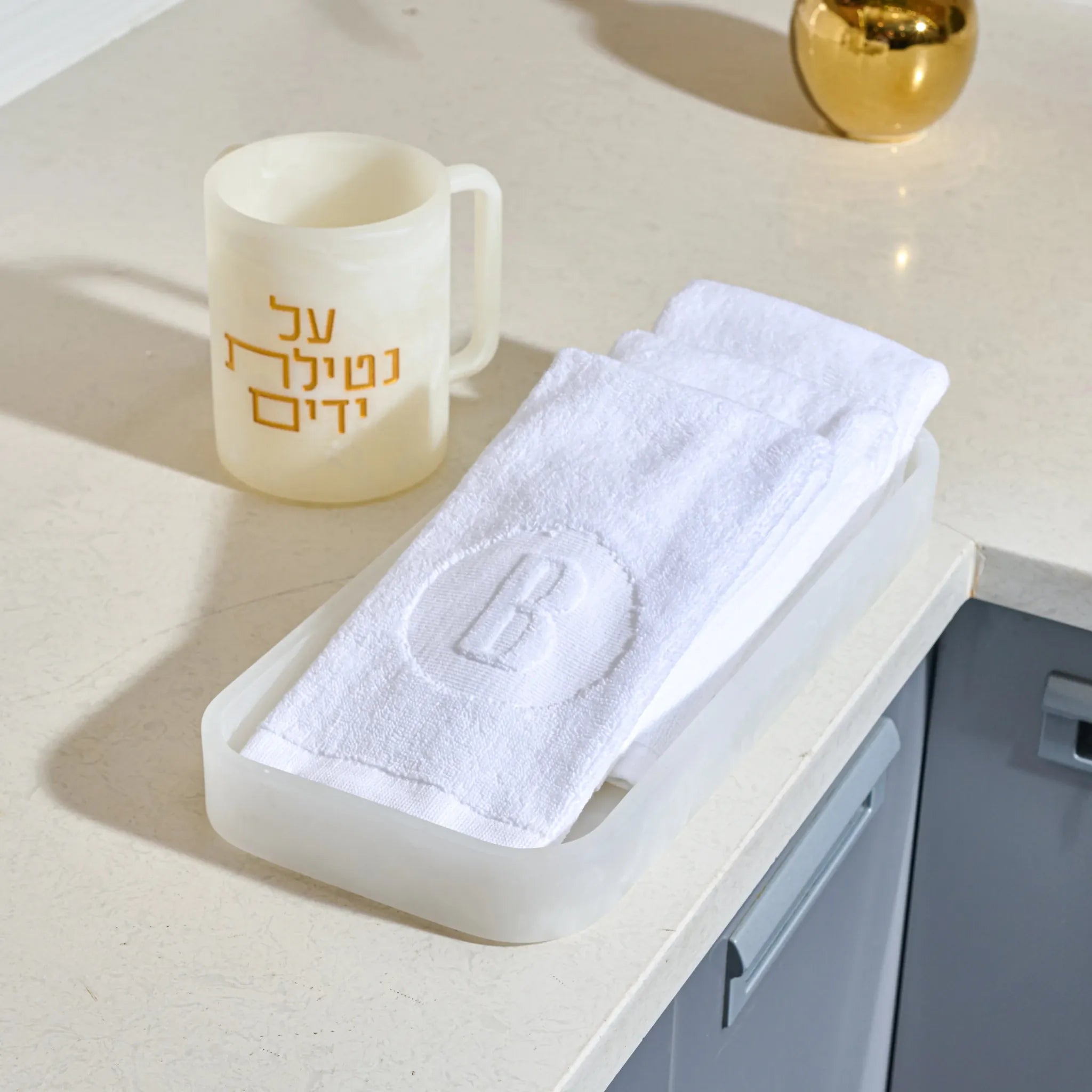 A white textured towel with an embroidered letter 'B' inside a circle, folded in a translucent white resin tray , set on a light beige countertop next to a cream-colored resin washing cup with gold Hebrew text.