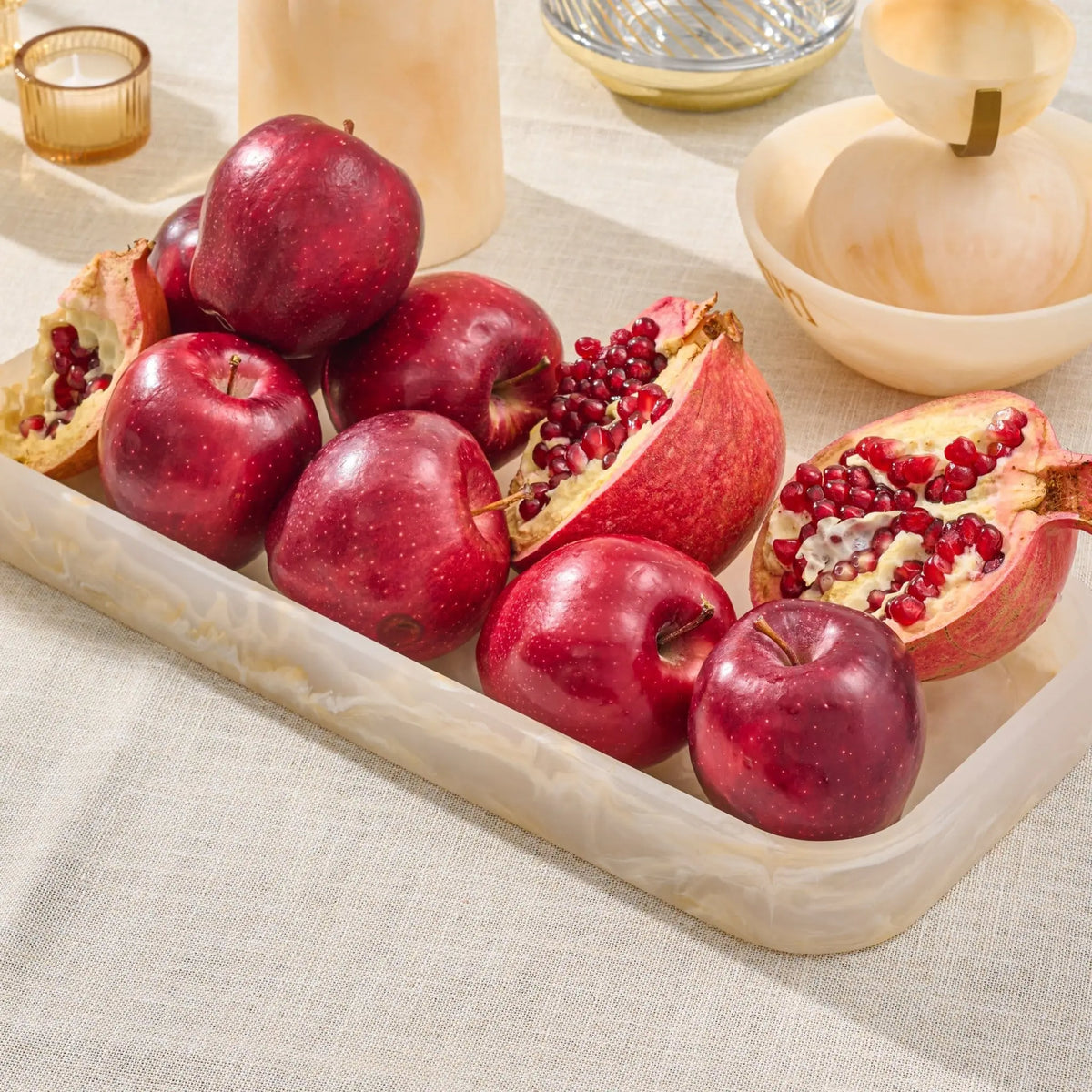 A rectangular resin tray with and cut open pomegranates, set on a light beige linen tablecloth with wooden bowls and candleholders in the background.