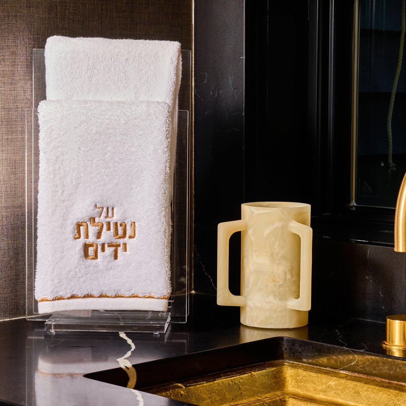 Cream marble-style resin washing cup with dual side handles, displayed beside a white embroidered “Al Netilat Yadayim” hand towel on a clear acrylic stand, set on a dark marble countertop.