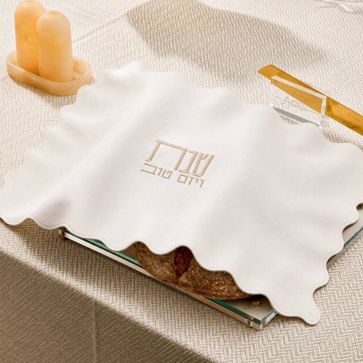 A white scalloped-edge cloth napkin with gold Hebrew lettering, draped over a book on a beige textured tablecloth.