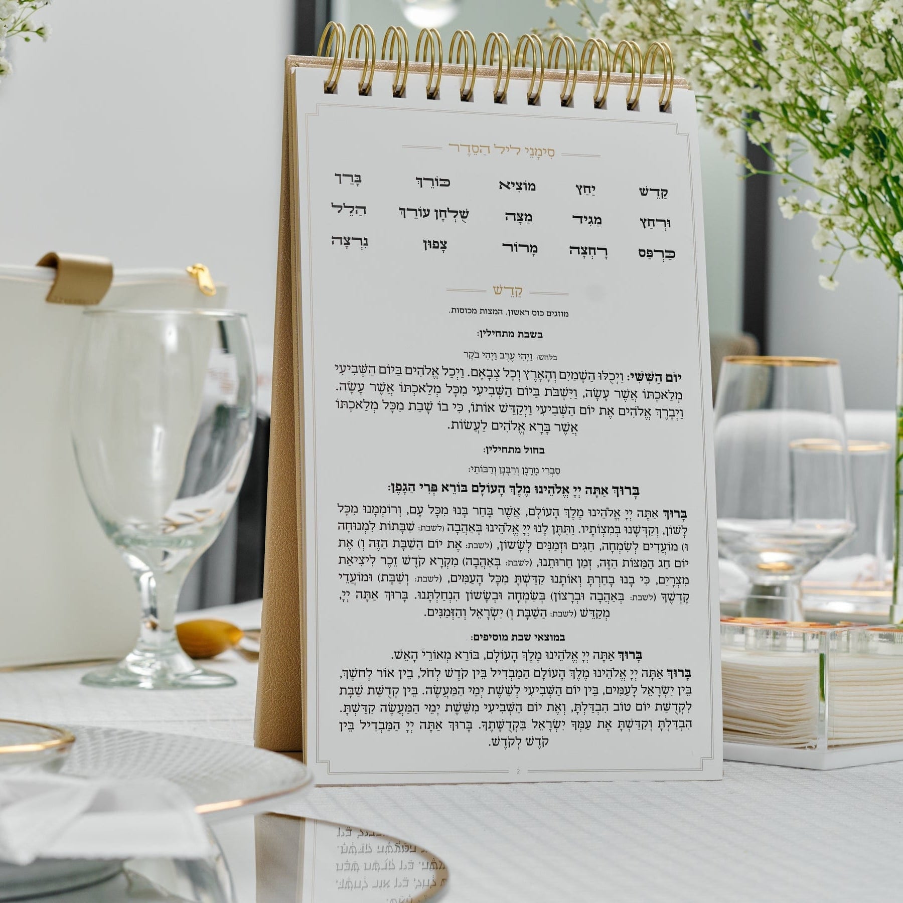 Gold Standing Leather Painted Haggadah on Shabbat table setting, gold flatware, glass plates, and vases filled with white baby’s breath flowers.