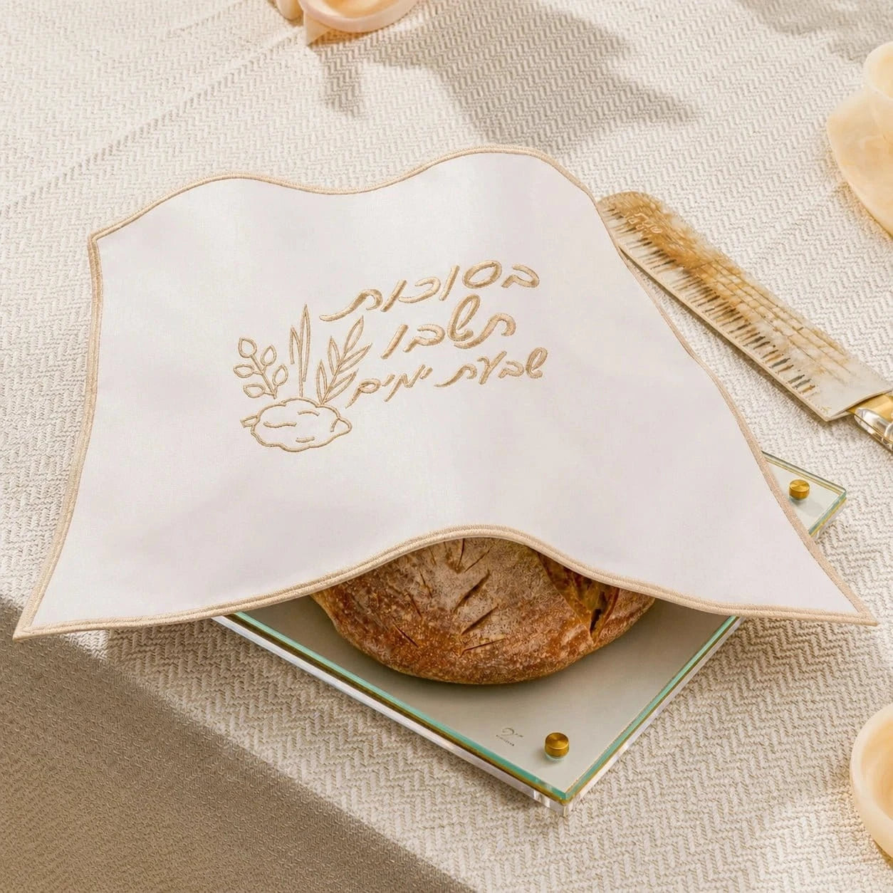 An off-white fabric challah cover with gold embroidered Hebrew text and a small botanical illustration, draped over a round loaf of bread on a glass plate. The scene is styled on a textured beige tablecloth with a gold knife in the background.