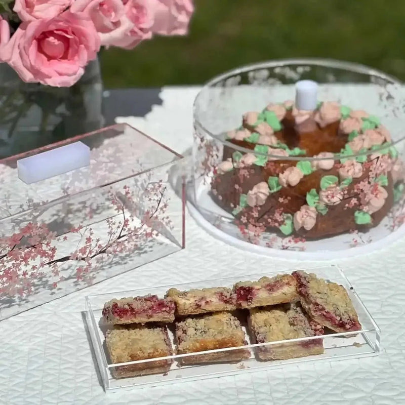 Clear Cherry Blossom cake dome with pink floral design, displayed with pastries and pink roses on white tabletop serveware.