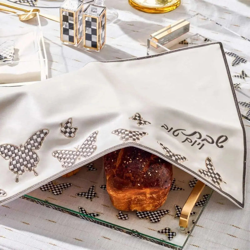 Onyx Butterfly Challah Cover in gold and silver leather displayed on a table with a glass of wine and challah bread during Shabbat dinner setup.