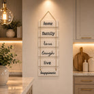 A vertical wooden sign with six white rectangular panels hanging by rope, each reading a positive word: home, family, love, laugh, live, happiness. The sign is displayed on a warm beige wall in a modern kitchen setting.
