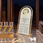 Chanukah brachos clock from Waterdale Collection, frosted Lucite in a gold arch frame with a working clock above the Chanukah brachos set up by night next to a window and menorah