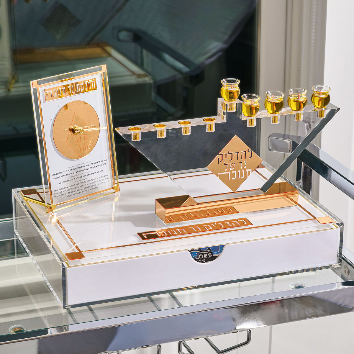 Gold Chanukah Brachos Clock styled on a glass shelf with a menorah tray and an oil jug, set for Hanukkah candle lighting