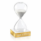 A clear glass hourglass for a chanuka timer with white sand, set on a polished gold rectangular base engraved with Hebrew text, shown on a clean white background.