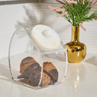 A clear acrylic and resin pastry container with a white lid, holding chocolate-dipped pastries, set on a light countertop next to a gold vase with pink flowers.