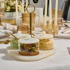 A styled tabletop setting with clear glass jars containing green olives and a beige spread, set on a resin tray with white lids and gold spoons. The scene includes amber glass candle holders with cream taper candles, ribbed amber glasses, and a white tablecloth in a bright, elegant dining environment.