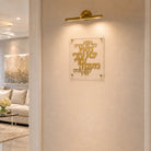 Gold Hebrew letter wall art mounted on a beige textured wall, illuminated by a gold wall sconce. The acrylic plaque is shown in a modern, elegant living room setting.