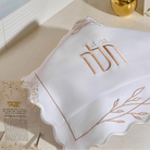 A white  linendecorative challah cover with a scalloped edge and a rose gold embroidered design featuring Hebrew text and leafy accents, set against a plain white background.