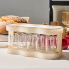 A cream-resin shot glass holder with gold trim, holding multiple small clear shot glasses, styled on a table with bread and holiday decor in the background.