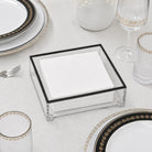 A clear square napkin holder with a black frame, filled with white folded napkins, styled on a white tablecloth with gold-rimmed plates, glasses, and a fork.
