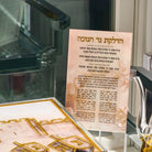 A printed Hebrew brachos card displayed on a clear stand, with a decorative gold-toned plaque or box in the foreground and a blurred indoor setting behind it.