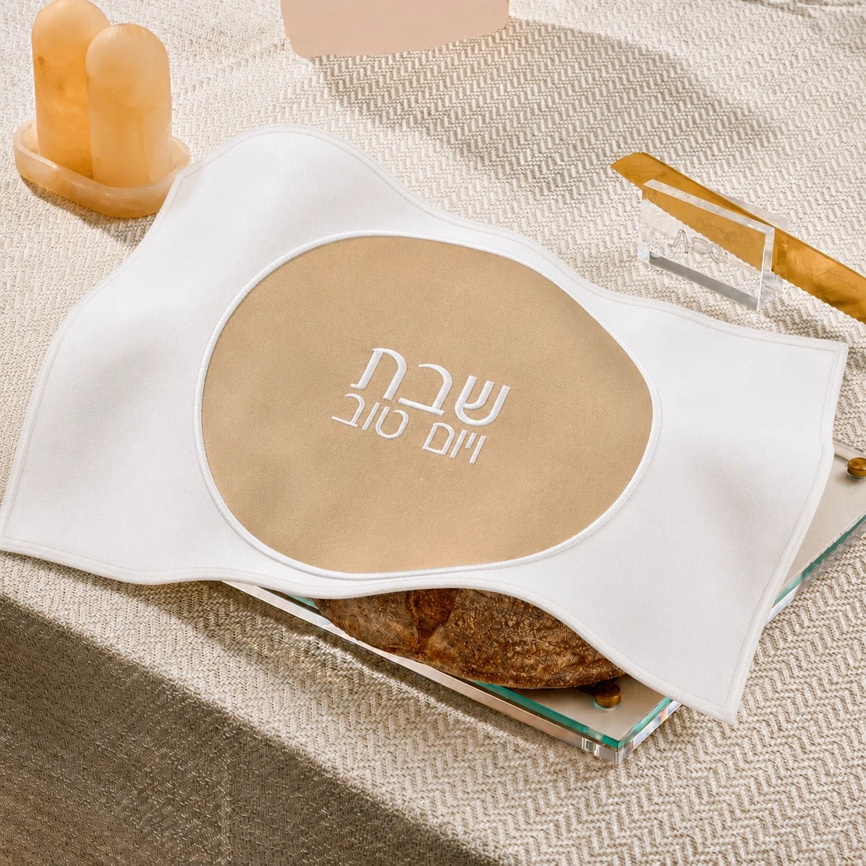 A white fabricchallah  cover with a large beige oval center printed with white Hebrew  text, styled on a textured beige surface with candles and a small dish in the background.