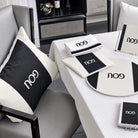 A styled interior scene showing coordinated black-and-whitePESACH SET  including a square throw pillow, folded napkins, placemat, sign, and notebook, arranged on a white tablecloth in a modern room.