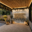 Outdoor patio area with a large stone and wood wall fountain mounted on the back wall, under a slatted wood ceiling with warm lighting and hanging greenery. The space includes a concrete floor, lanterns with candles, potted plants, and a wooden fence in the background.