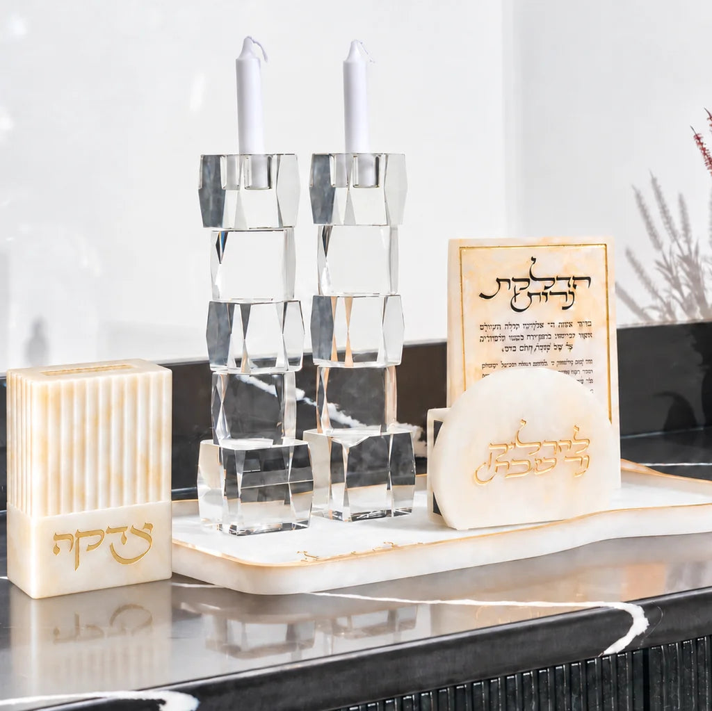 Gold resin tzedaka box beside clear faceted Shabbat candlesticks and a Hadlakas Neiros card on a marbled tray