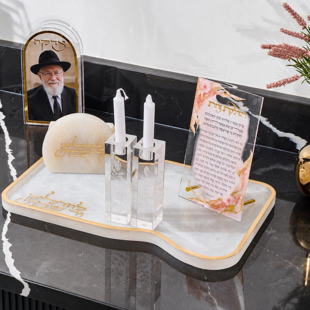 A decorative white and gold resin tray on a black marble surface, holding a small framed portrait, two clear candle holders with white candles, a cream stone, and a printed card. A gold vase with pink flowers sits to the right.