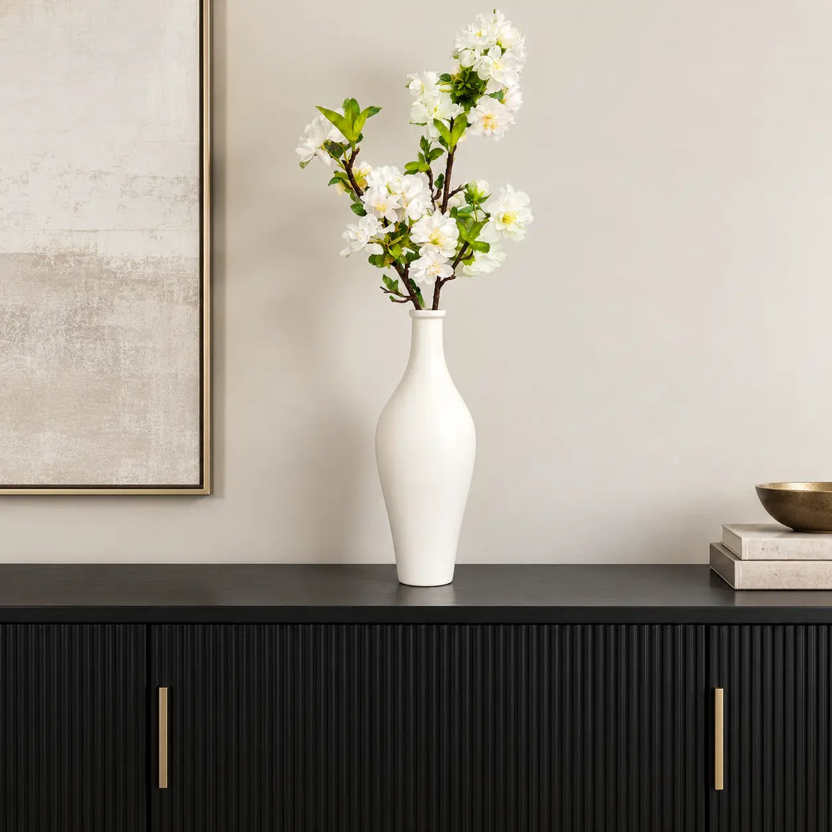 Tall white ceramic vase with white faux blossom branches styled on a black fluted sideboard under a framed canvas