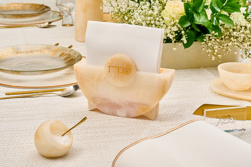 Elegant Shabbat table setting featuring a marble-look napkin holder with Hebrew “Shabbat” inscription, surrounded by gold-rimmed plates, floral arrangement, and neutral-toned tableware. | Waterdale Collection