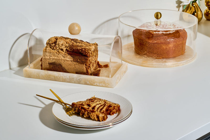 Elegant acrylic cake dome and marble-look base set displaying baked goods, including a loaf cake and round cake, styled with gold-accented serveware for a modern dessert presentation. | Waterdale Collection