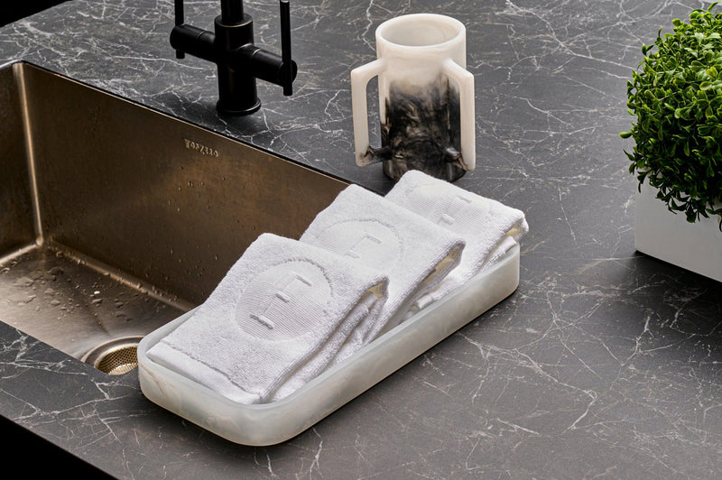 Modern netilat yadayim set featuring a marble-look washing cup and matching tray with embroidered hand towels, styled on a dark stone countertop beside a sink. | Waterdale Collection