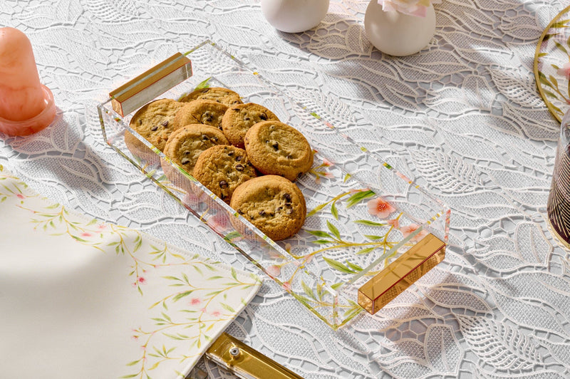 Clear acrylic serving tray with gold handles holding chocolate chip cookies, styled on an elegant lace tablecloth with floral accents for a refined dessert presentation. | Waterdale Collection