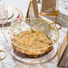 Floral bird matzah box with gold-tone metal frame holding round matzo, displayed on decorative table setting with place card and wine glass.