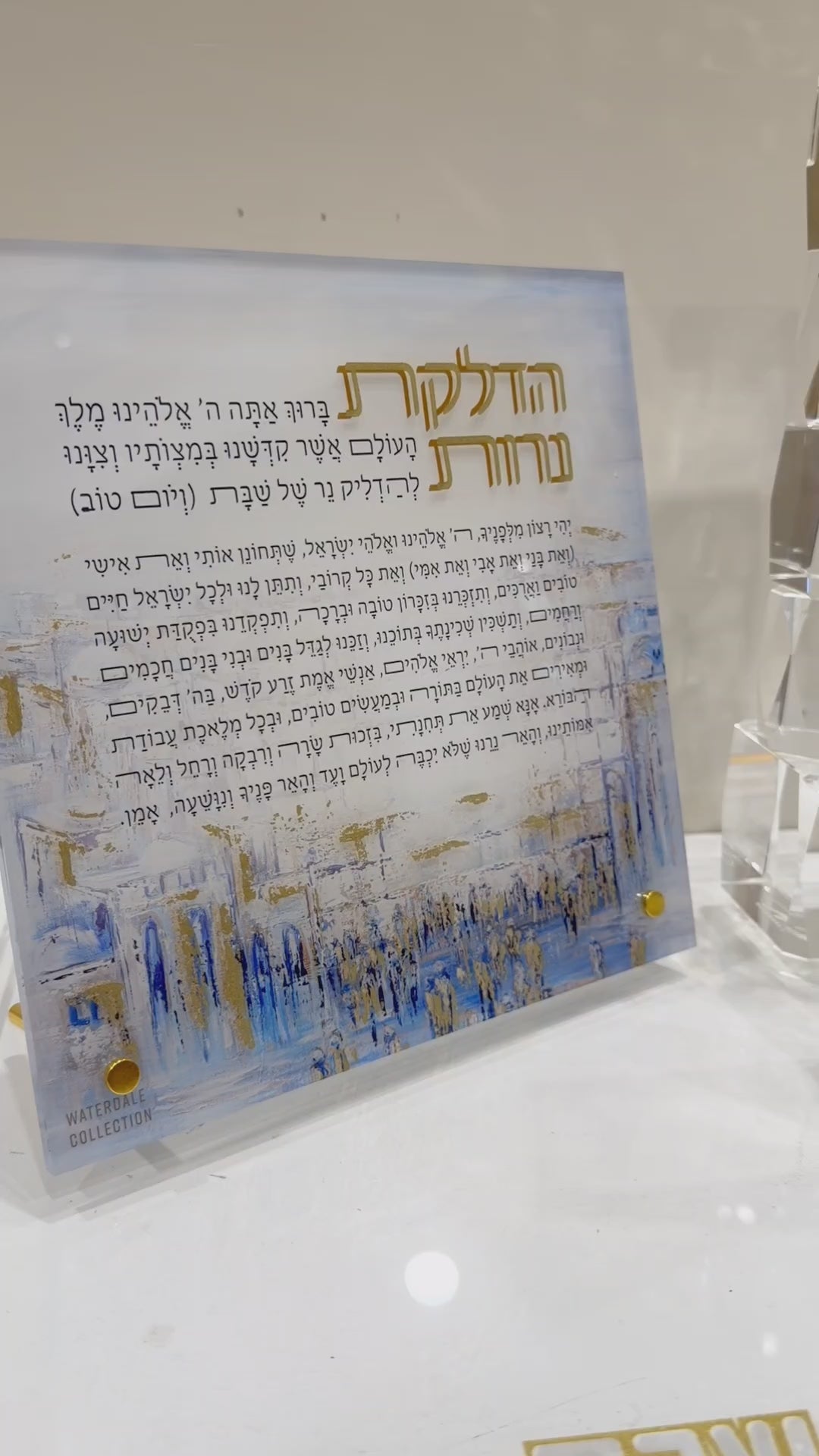 Coins being put into the Painted Yosefa Kosel Tzedakah Box featuring a serene blue-toned painting of the Kosel with gold “Tzedakah” lettering, encased in a clear lucite box with a sleek gold base.