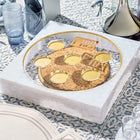 Pearl marble Seder plate with gold-rimmed circular wells and Hebrew text detail, displayed on patterned tablecloth with water glass and wine bottle.