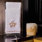 Ombre resin washing cup with gold “Al Netilas Yadayim” Hebrew lettering, displayed beside a white embroidered hand towel on a sleek black marble countertop near a modern sink.