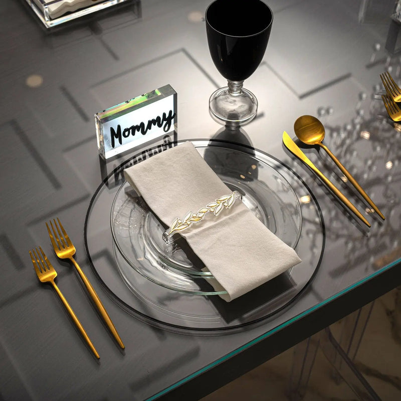 Black and gold tablescape set with clear plates, beige napkin, gold utensils, black wine glass, and place card for elegant dining.