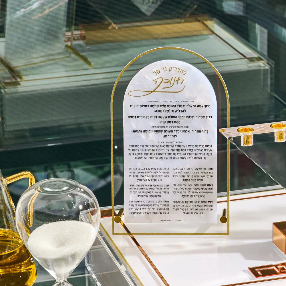 A gold-framed chanukah blessing card , displayed on a glass table alongside a glass of white wine and a glass of oil.