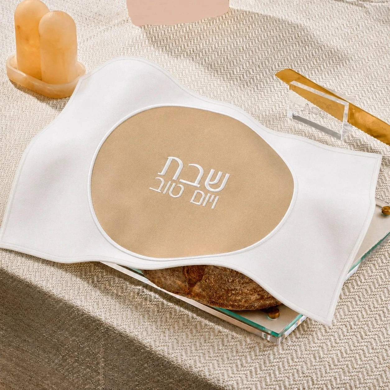 A white fabricchallah  cover with a large beige oval center printed with white Hebrew  text, styled on a textured beige surface with candles and a small dish in the background.