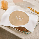 A white fabricchallah  cover with a large beige oval center printed with white Hebrew  text, styled on a textured beige surface with candles and a small dish in the background.