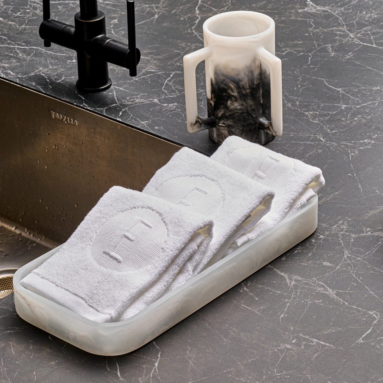 White circle initial embossed finger towels folded in a rectangular marble-look resin tray, displayed on a dark stone countertop beside a black-and-white marble resin washing cup and modern matte black faucet.