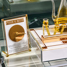 A gold-framed acrylic display stand holding a printed card with Hebrew text and a circular gold illustration, placed on a glass shelf beside a gold decorative tray and a glass vessel filled with golden liquid.