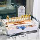 A white and gold Hanukkah menorah set with nine small gold cups filled with golden liquid, displayed on a glass cart. The set includes a white drawer base with compartments holding packaged candes glassesss....