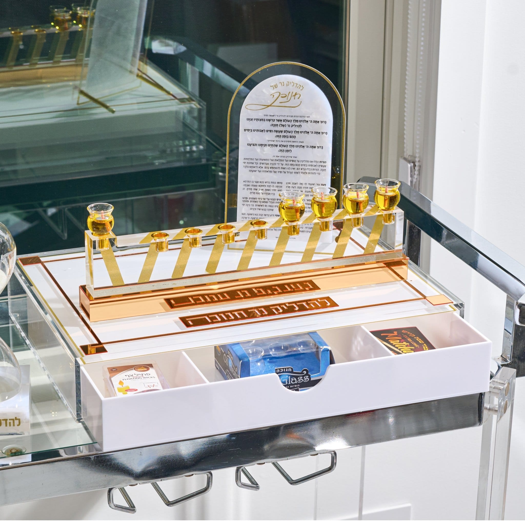 A white and gold Hanukkah menorah set with nine small gold cups filled with golden liquid, displayed on a glass cart. The set includes a white drawer base with compartments holding packaged candes glassesss....