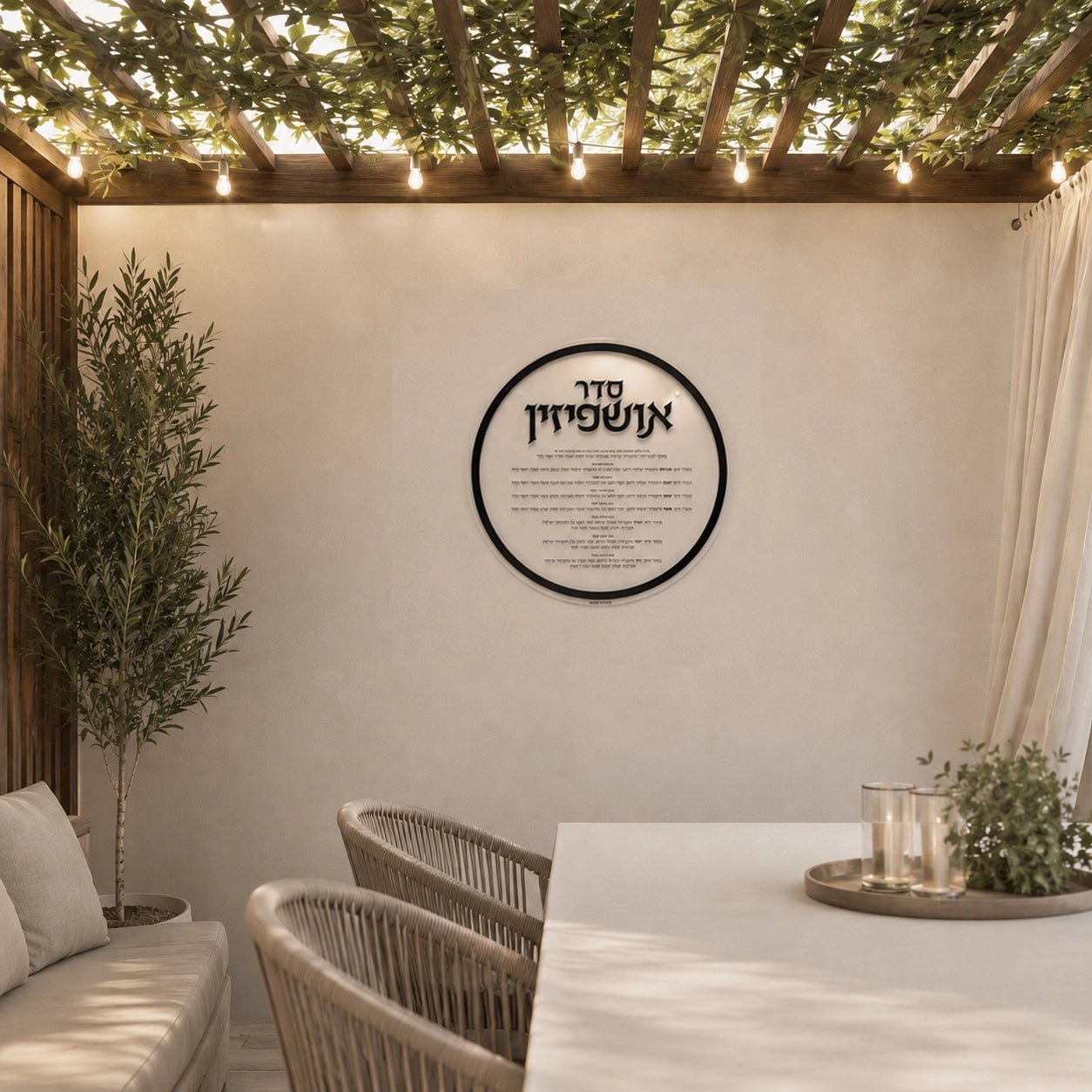 Round clear Lucite Ushpizin sukkah decoration hanging in a modern succah with a Sukkot table, chairs, and potted plants.