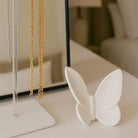 Matte white ceramic or resin butterfly sculpture/figurine displayed on a white bedside table next to a clear acrylic and metal jewelry stand holding gold and silver chain necklaces - Waterdale Collection