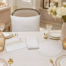 A styled dining table setting featuring a white embroidered pesah set cushion, matching white napkins, a white placemat, and gold-accented tableware in a bright, elegant room.