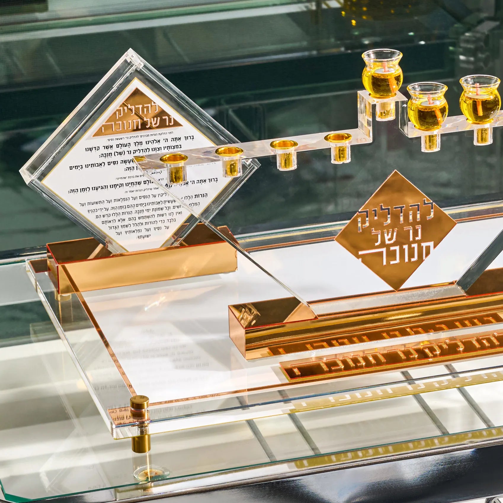 A clear acrylicHanukkah menorah set with a diamond-shaped Hebrew inscription plaque and a gold-toned base, photographed on a reflective glass surface.