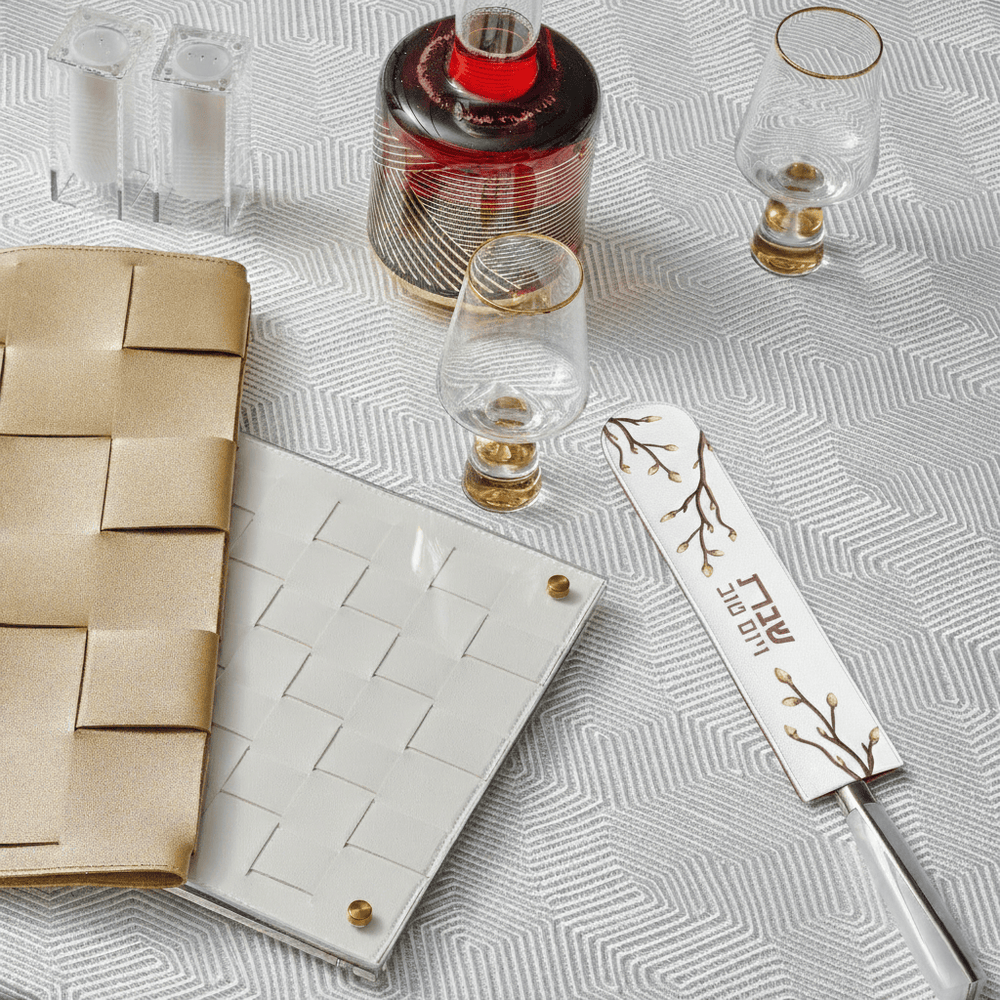 White embroidered leaf knife blade cover with a gold branch design and Hebrew inscription, resting on a geometric white tablecloth next to a challah knife - Waterdale Collection