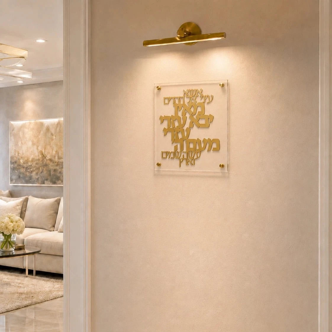 Gold Hebrew letter wall art mounted on a beige textured wall, illuminated by a gold wall sconce. The acrylic plaque is shown in a modern, elegant living room setting.